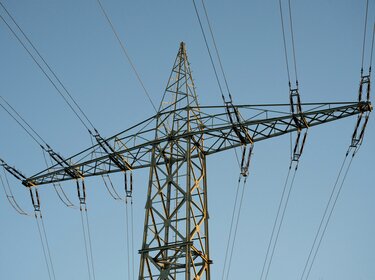 Hybride Angriffe: Sicherheitstechnische Infrastruktur ertüchtigen Ein hoher Strommast aus Stahlgitter gegen einen blauen Himmel.