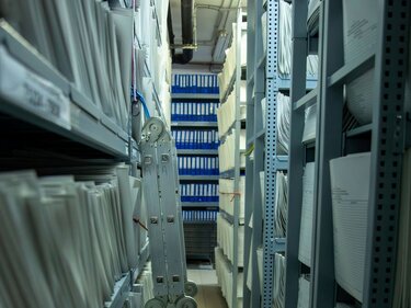 Blick in einen Kellerraum, in dem sich die Last der Bürokratie in Form voller Bücherregale und überquellender Ordner zeigt