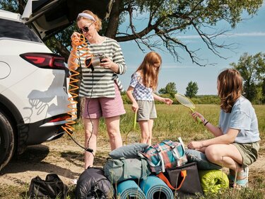 Eine Mutter und ihre Töchter stehen am Kofferraum eines weißen Autos vor einer Menge Gepäck