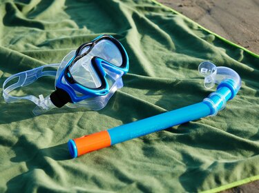Eine Tauchmake mit blauem Rand und eine blaue Schnorchel mit orangefarbener Spitze liegen auf einem grünen Handtuch. 