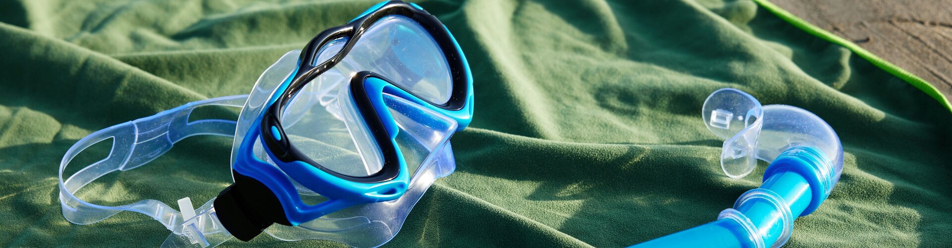Sicher unter Wasser: Darauf kommt es bei Tauchmaske und Schnorchel an Eine Tauchmake mit blauem Rand und eine blaue Schnorchel mit orangefarbener Spitze liegen auf einem grünen Handtuch.