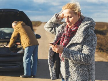 Sicher durch die Ferien: Tipps für Stau, Panne und Unfall Eine Frau steht mit besorgtem Blick am Straßenrand und schaut auf ihr Smartphone, während sie sich mit der anderen Hand an den Kopf fasst. Im Hintergrund versucht eine weitere Person bei geöffneter Motorhaube ein liegengebliebenes Auto zu reparieren. Die Szene spielt auf einer abgelegenen Landstraße unter bewölktem Himmel.