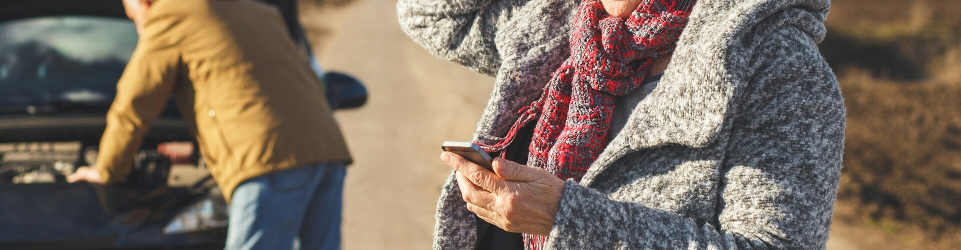 Sicher durch die Ferien: Tipps für Stau, Panne und Unfall Eine Frau steht mit besorgtem Blick am Straßenrand und schaut auf ihr Smartphone, während sie sich mit der anderen Hand an den Kopf fasst. Im Hintergrund versucht eine weitere Person bei geöffneter Motorhaube ein liegengebliebenes Auto zu reparieren. Die Szene spielt auf einer abgelegenen Landstraße unter bewölktem Himmel.