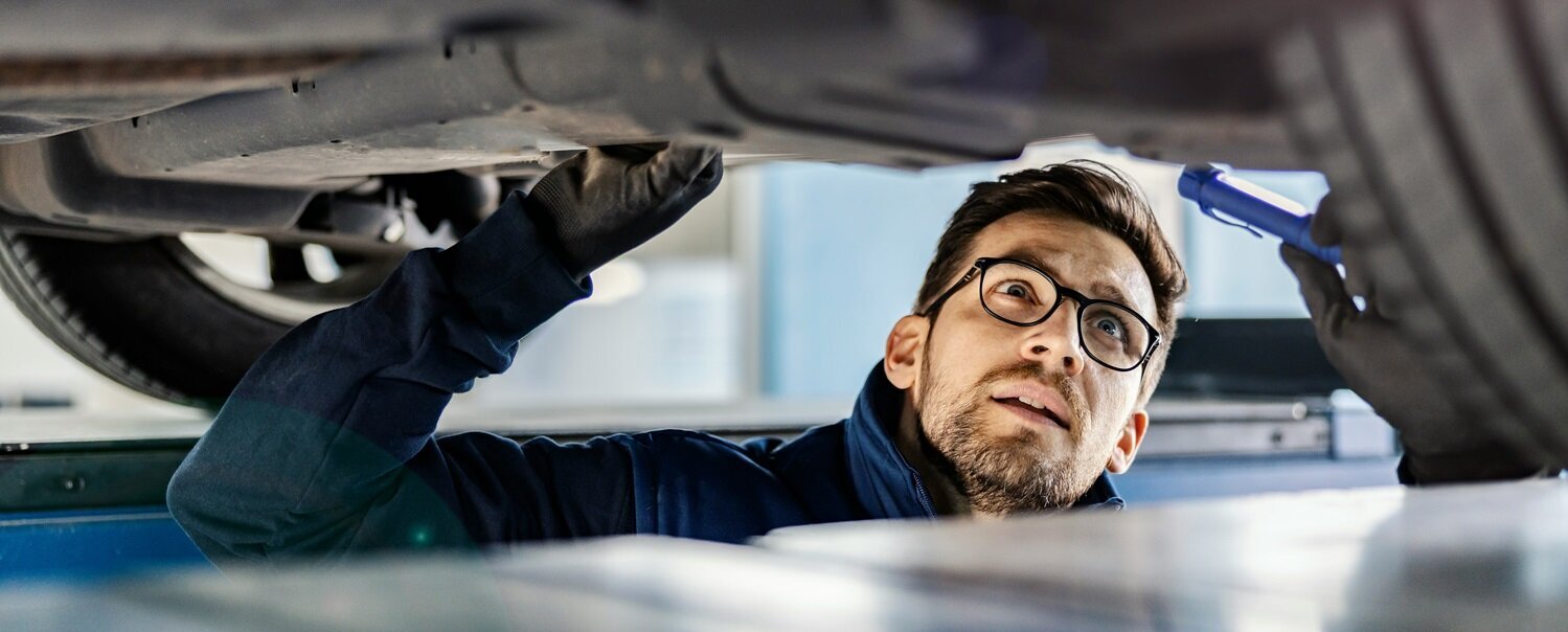 Ein Mann steht in der Montagegrube unter einem Pkw und leichtet die Unterseite des Fahrzeugs mit einer Taschenlampe an