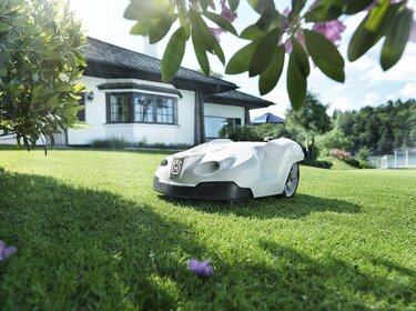 weißer Rasenmäher-Roboter fährt auf einer Wiese vor einem weißen Einfamilienhaus