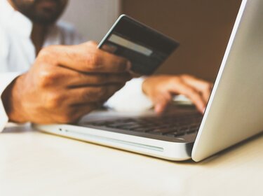 FFP2- & OP-Masken online kaufen Mann hält Kreditkarte in einer Hand und tippt die Informationen am Laptop ein