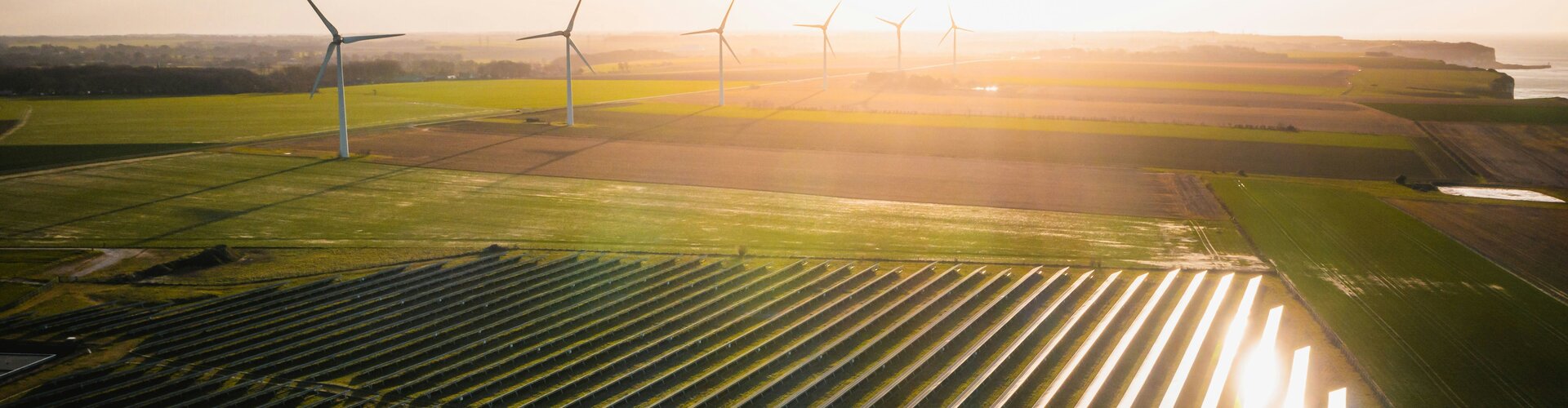 TÜV-Verband-Merkblatt: Basisrichtlinie zur Ökostromzertifizierung aktualisiert Luftaufnahme eines Solarparks und mehrerer Windräder auf weitläufigen Feldern im warmen Licht der tief stehenden Sonne.