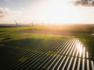 TÜV-Verband-Merkblatt: Basisrichtlinie zur Ökostromzertifizierung aktualisiert Luftaufnahme eines Solarparks und mehrerer Windräder auf weitläufigen Feldern im warmen Licht der tief stehenden Sonne.