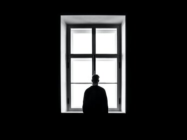 Mann steht in dunklem Zimmer vor hellstrahlendem Fenster und schaut hinaus