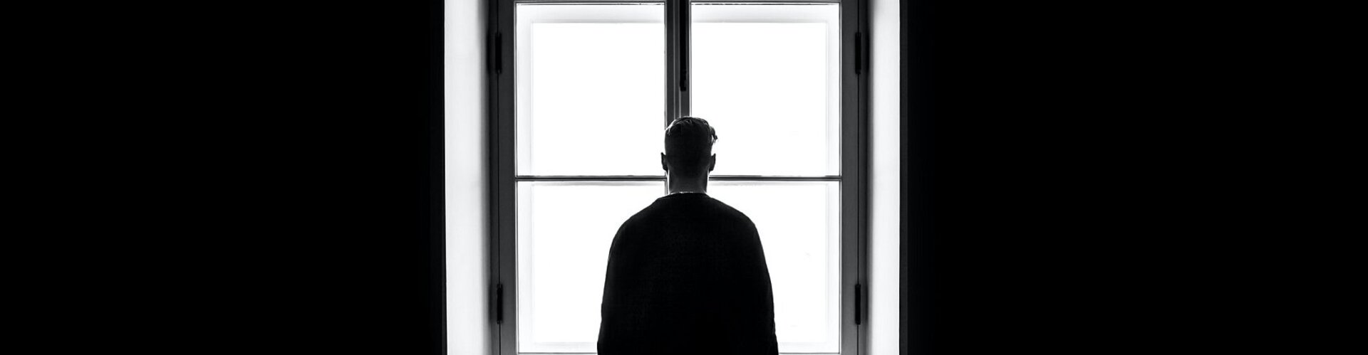 Mann steht in dunklem Zimmer vor hellstrahlendem Fenster und schaut hinaus