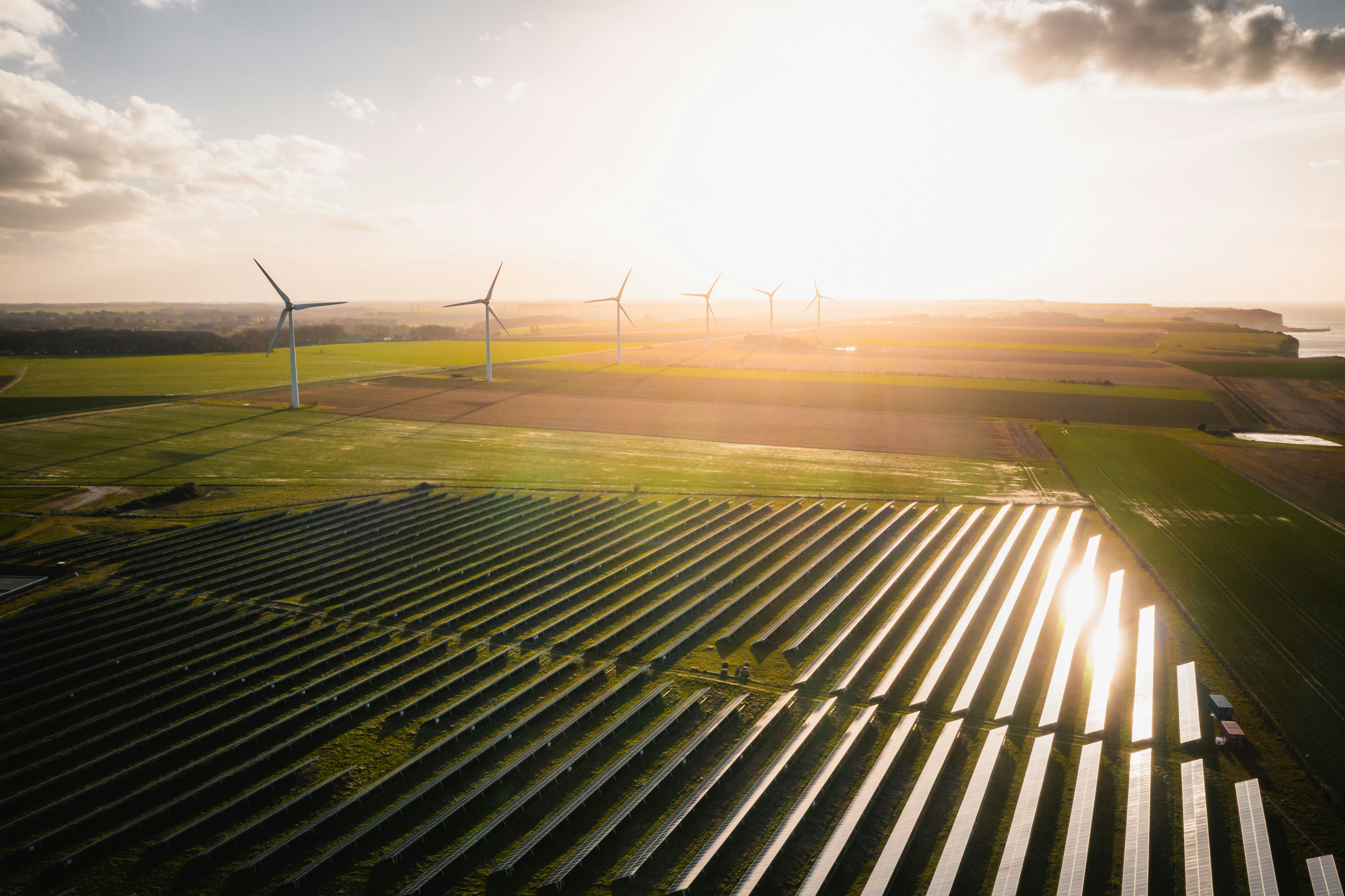 Luftaufnahme eines Solarparks und mehrerer Windräder auf weitläufigen Feldern im warmen Licht der tief stehenden Sonne.