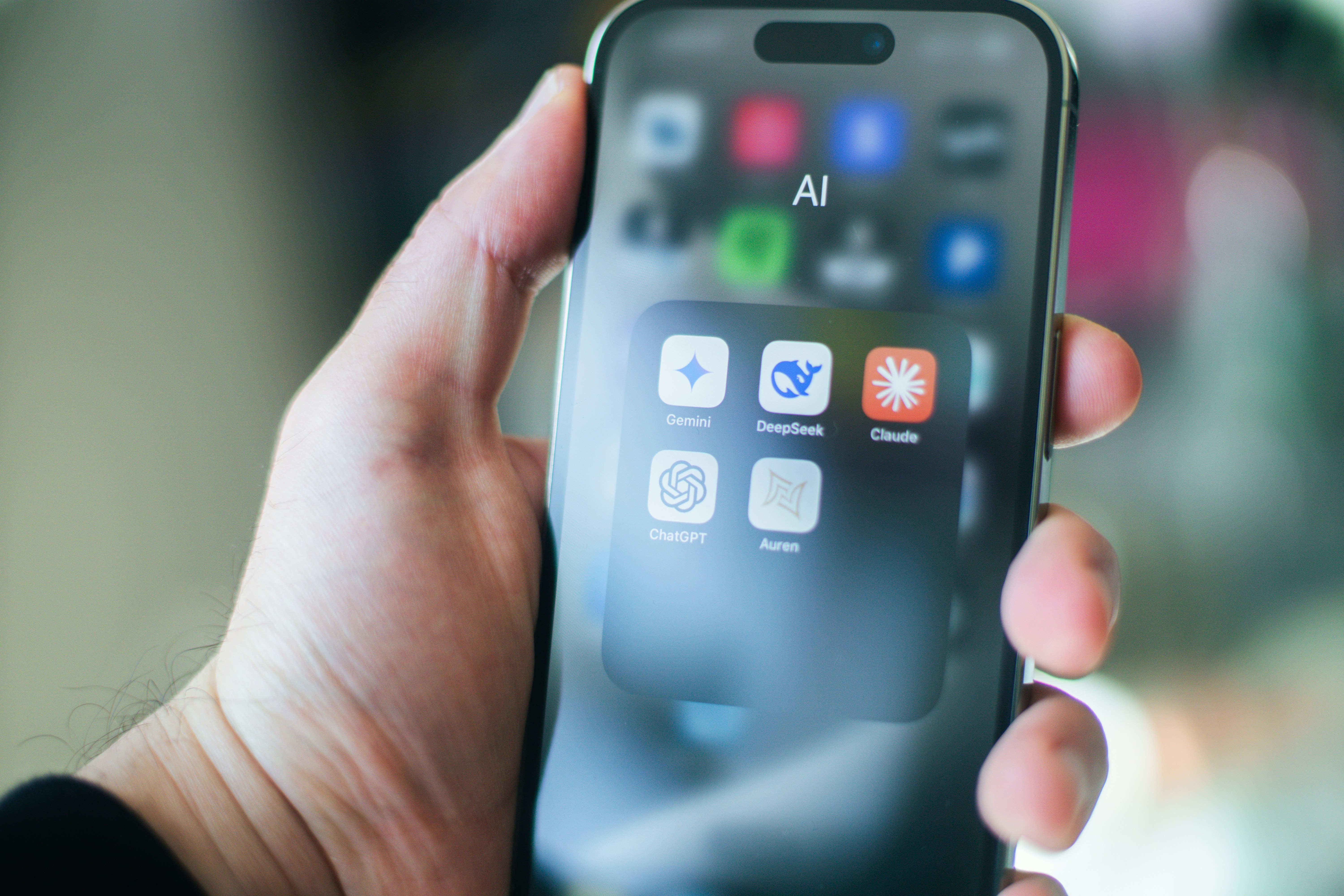 In der Hand hält jemand ein Handy. Auf dem Bildschirm ist ein Ordner geöffnet in dem sich verschiedene KI-Apps befinden. Beispielsweise Claude oder Chatgpt.